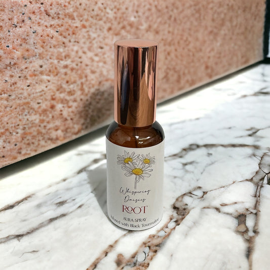 A glass bottle of 'Root - Tourmaline Black - Aura Spray' with a copper-colored cap, standing on a marble surface. The label features a floral design and the text 'Whispering Daisies ROOT AURA SPRAY'.